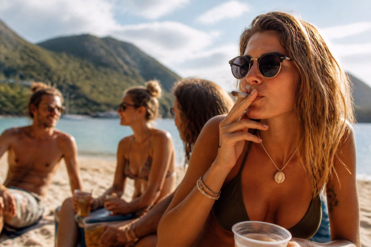 Woman in sunglasses smoking a joint on a tropical beach with friends holding drinks in the background