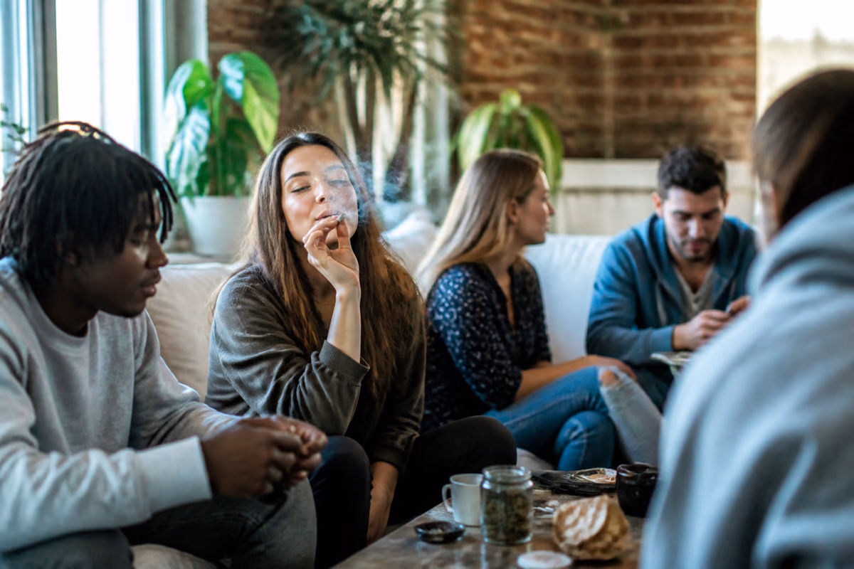 Young adults on a sofa as a woman smokes a joint, with a jar of cannabis buds on the coffee table